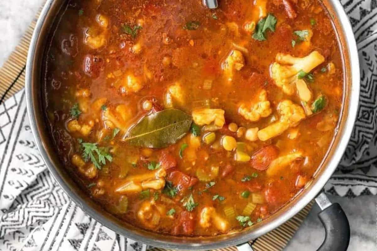 A pot filled with a hearty stew featuring pieces of cauliflower, beans, tomatoes, and various vegetables in a rich, tomato-based broth. A bay leaf floats on top, and the stew is garnished with fresh herbs. The pot is placed on a patterned cloth.