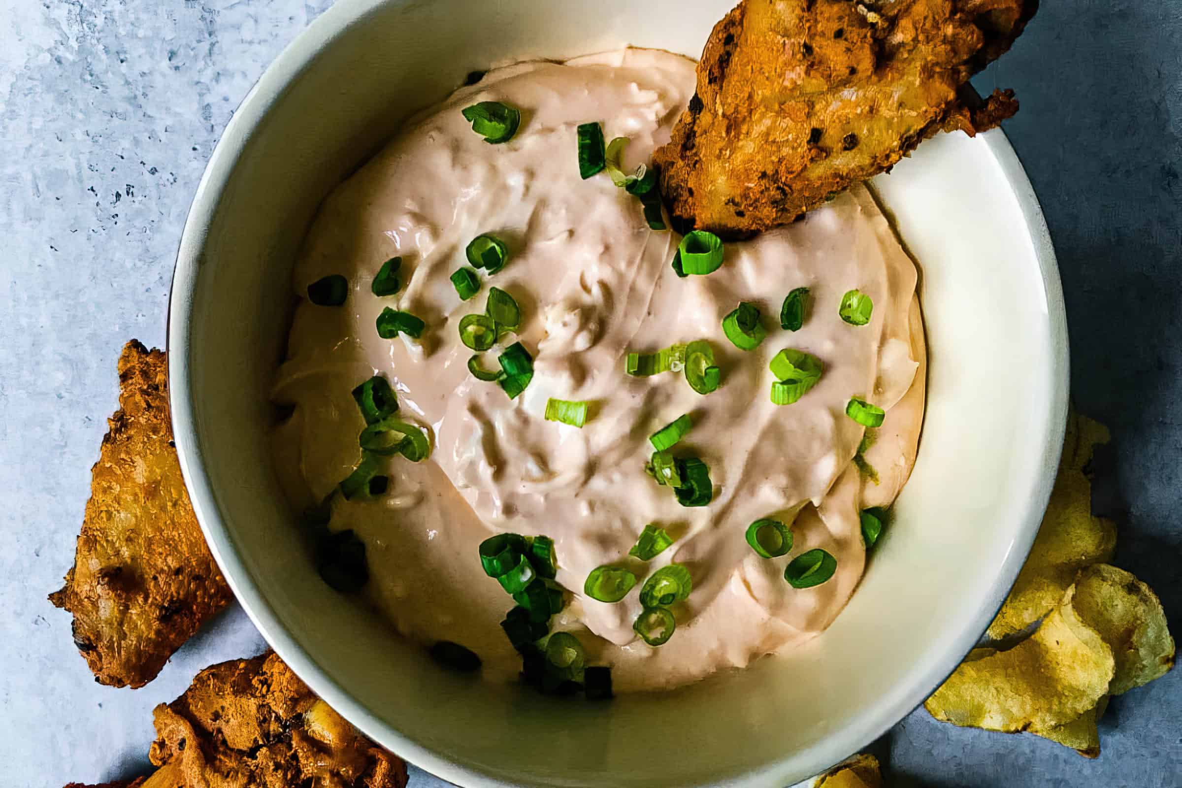 A white bowl filled with creamy dip topped with chopped green onions. A piece of fried food is partially dipped into the sauce, with additional fried items and chips placed around the bowl on a light surface.