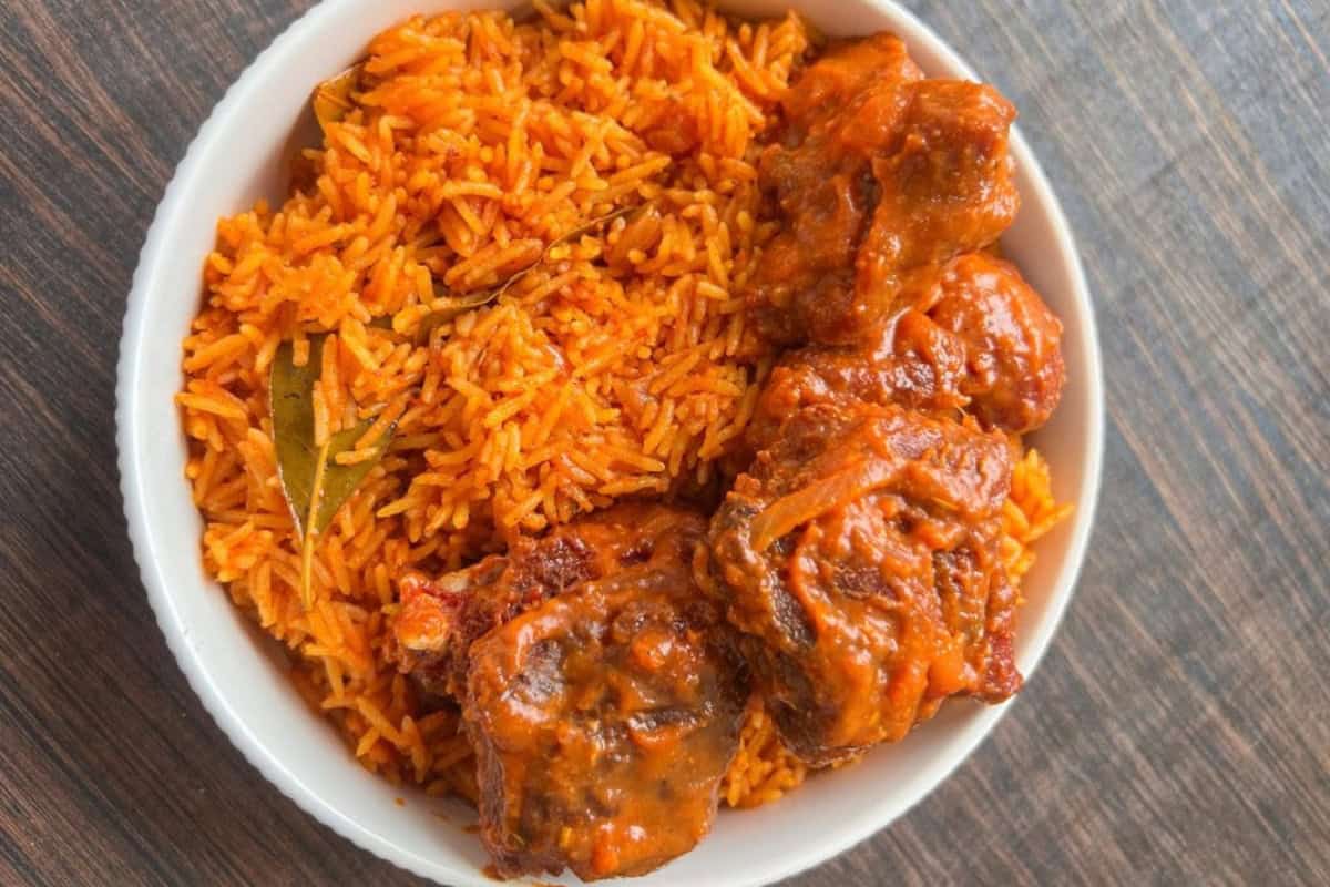 A white bowl filled with orange-colored jollof rice and three pieces of stewed meat is placed on a wooden table. The rice is garnished with bay leaves. The hearty dish has a vibrant, appetizing appearance.