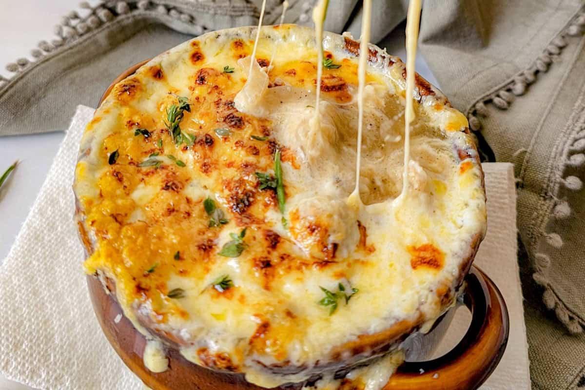 A close-up of a bowl of French onion soup topped with melted, browned cheese being stretched with a spoon. The dish is garnished with chopped herbs and placed on a white napkin. In the background, a green cloth with a fringed edge is partially visible.