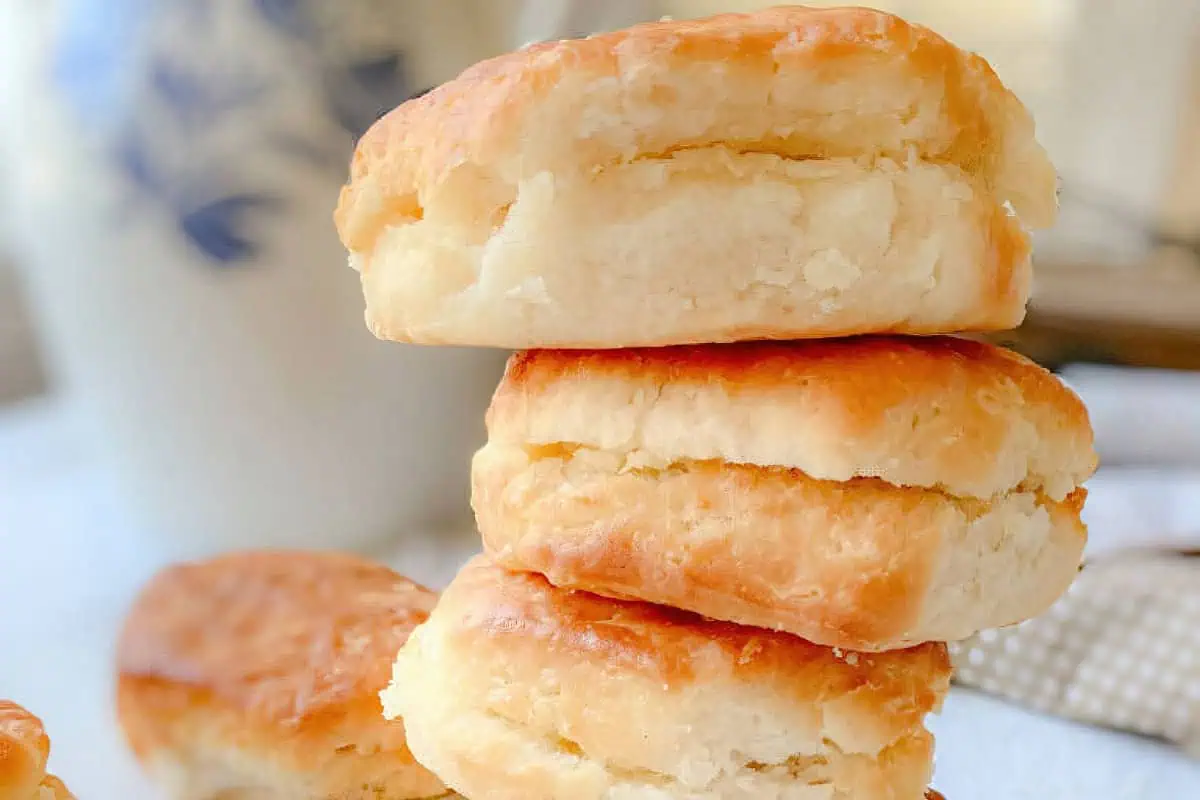 A stack of flaky, golden-brown biscuits is piled high on a table. The layers are visible, showing the light and fluffy texture inside. In the background, part of a white and blue patterned mug is slightly blurred.