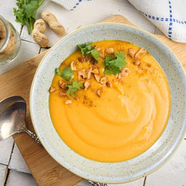 A bowl of creamy orange soup with chopped peanuts and cilantro on top, set on a wooden board with whole peanuts, a spoon, and a towel.