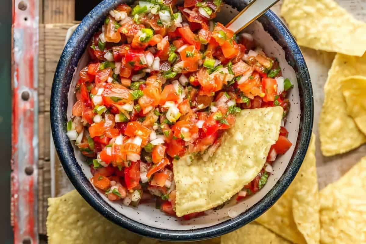 A close-up of a bowl of fresh salsa, filled with diced tomatoes, onions, cilantro, and jalapeños. A spoon is resting in the bowl, and several tortilla chips are placed around and in the salsa, ready for dipping.