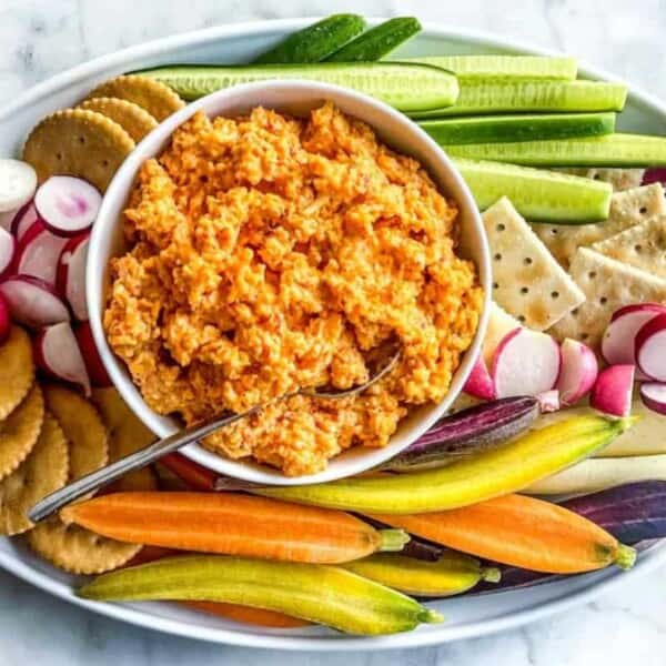 A platter with a bowl of orange dip in the center, surrounded by sliced cucumbers, radishes, colorful carrots, round crackers, and square crackers, all arranged neatly on a white oval serving dish. A spoon rests in the dip. The background is a marble surface.