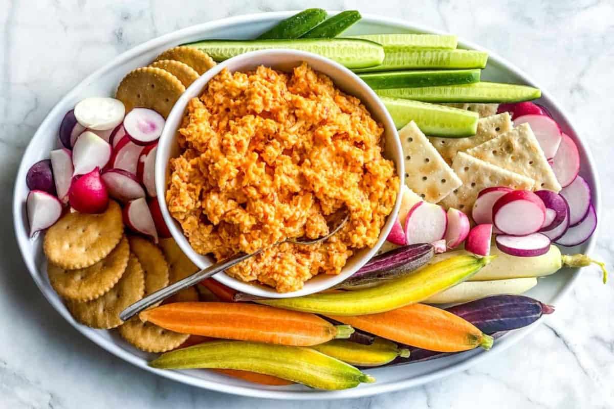 A platter with a bowl of orange dip in the center, surrounded by sliced cucumbers, radishes, colorful carrots, round crackers, and square crackers, all arranged neatly on a white oval serving dish. A spoon rests in the dip. The background is a marble surface.