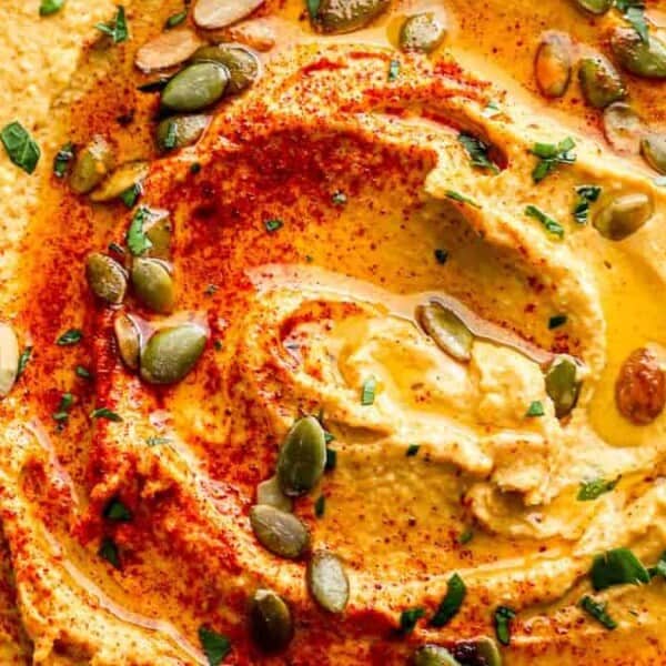 A bowl of hummus with paprika, olive oil, pumpkin seeds, and chopped parsley on top.