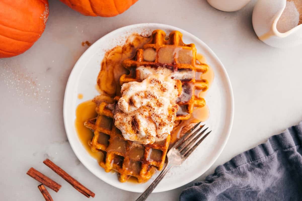A white plate with two pumpkin waffles topped with whipped cream and a dusting of cinnamon. Syrup is drizzled over the waffles. A fork rests on the plate's edge. Surrounding the plate are a couple of whole pumpkins, cinnamon sticks, and a blue napkin.