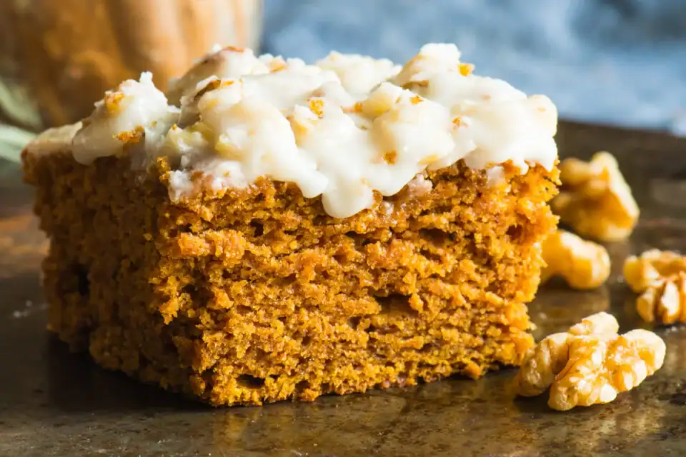 Close-up of a single slice of moist pumpkin cake topped with creamy frosting and chopped nuts, likely walnuts, displayed on a rustic metal surface. Additional walnut pieces are scattered around the slice, with a light blue background softly blurred.