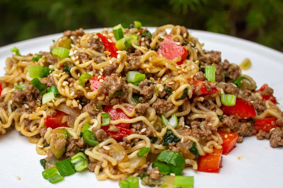 A close-up of a plate of ramen noodles mixed with ground beef, chopped green onions, red bell peppers, and sprinkled with sesame seeds. The dish looks colorful and garnished with fresh vegetables, creating an appetizing appearance.