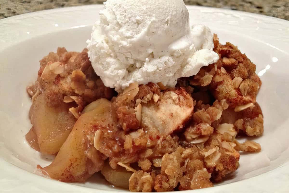 A bowl of apple crisp topped with a scoop of vanilla ice cream. The apple crisp is golden brown with visible oats and apple slices, creating a textured, crumbly appearance. The ice cream is melting slightly over the warm dessert, adding a creamy contrast.