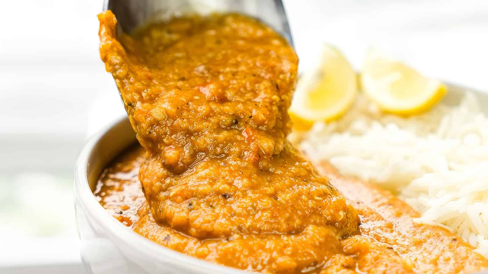 A ladle pours thick, spiced lentil curry into a bowl with a side of lemon wedges and rice.