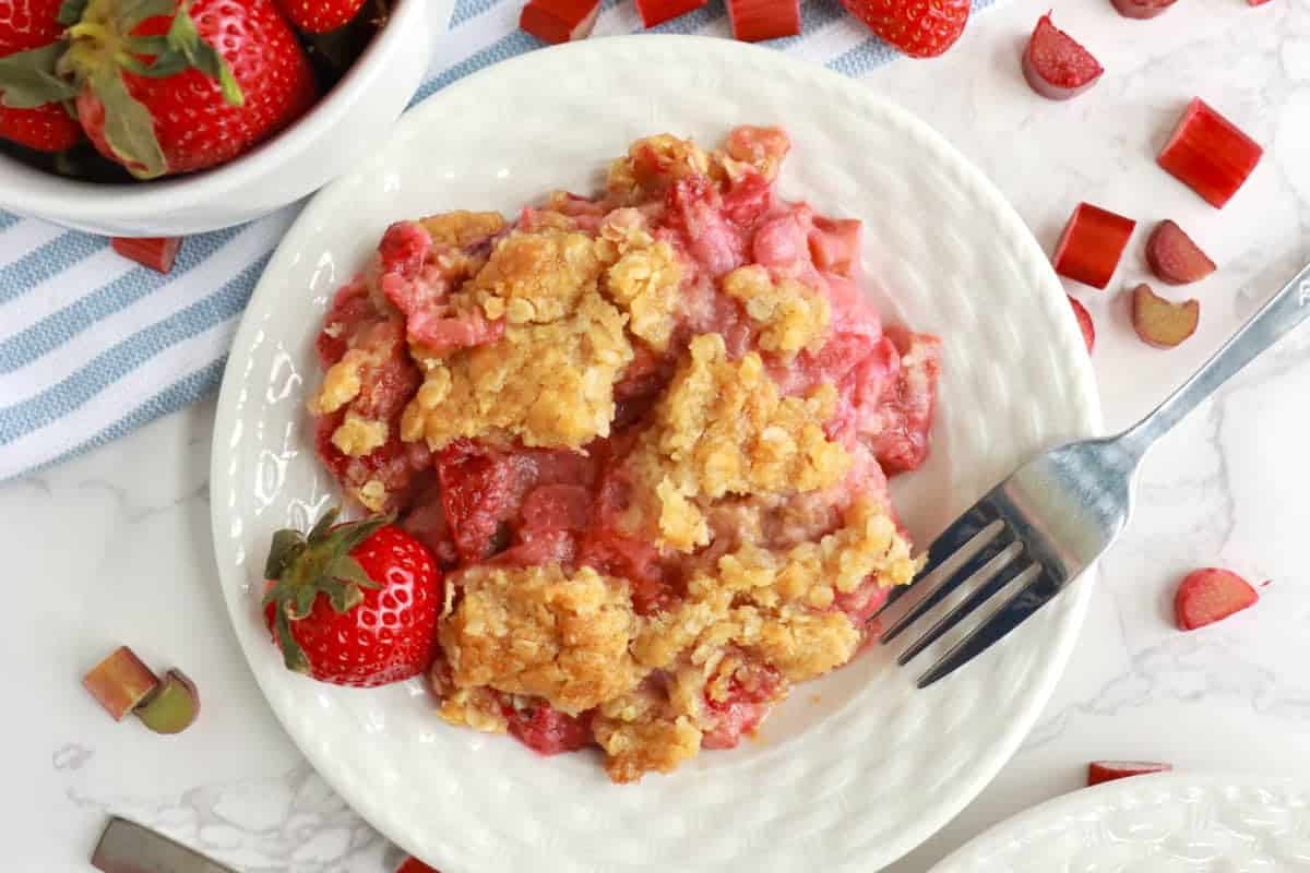 A plate of strawberry rhubarb crisp, garnished with a fresh strawberry, sits on a white marble countertop. The crisp features a golden, crumbly topping over the pinkish filling. A fork rests on the plate and scattered rhubarb pieces surround it.