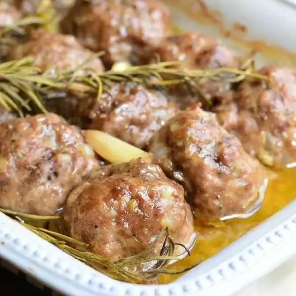 A close-up of a casserole dish filled with browned meatballs garnished with sprigs of rosemary and garlic cloves. The meatballs are sitting in a light, flavorful broth. The dish has a white, patterned edge.
