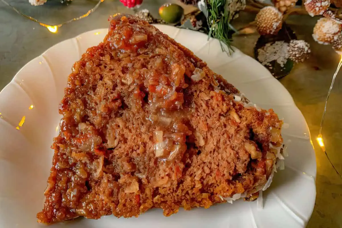 A piece of moist, spiced carrot cake sits on a white plate. It has a crumbly texture, filled with chunks of carrot and nuts and topped with a caramel-like glaze. The plate is set against a background with festive holiday décor and soft, warm lighting.