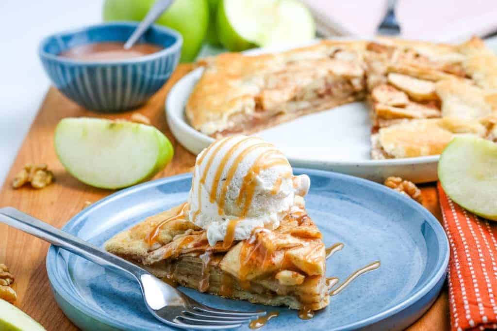 A slice of apple pie with a scoop of vanilla ice cream drizzled with caramel sauce on a blue plate with a fork. The remaining pie and a bowl of caramel sauce are in the background with green apple slices and a red cloth napkin.