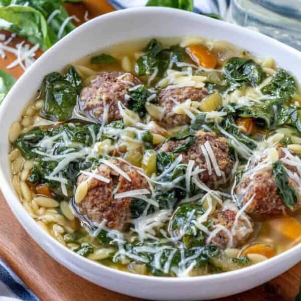 A bowl of Italian wedding soup filled with vibrant green spinach, meatballs, diced carrots, celery, and orzo pasta, topped with grated Parmesan cheese. The soup is served in a white bowl on a wooden surface, surrounded by fresh spinach leaves.