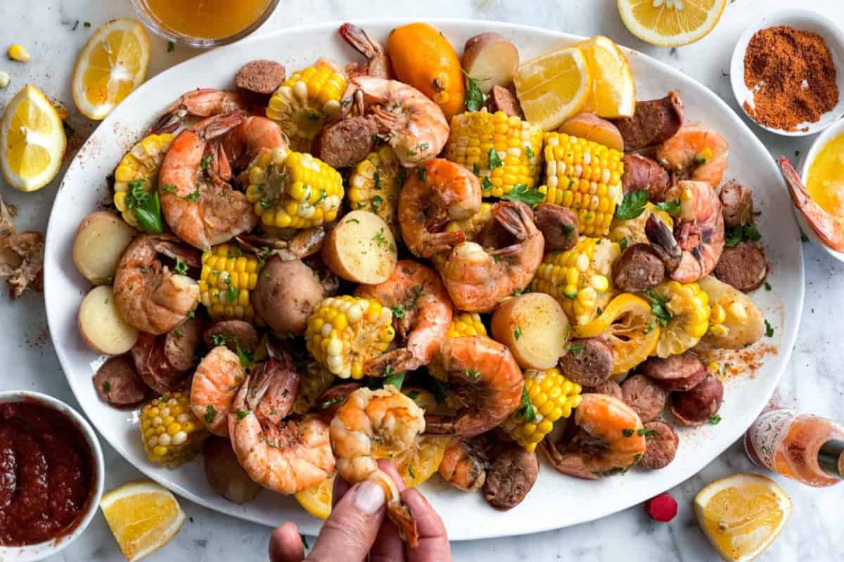A large white platter filled with a seafood boil, featuring shrimp, sausage, corn on the cob, and boiled potatoes, garnished with fresh herbs. Lemon wedges and dipping sauces surround the platter. A hand is taking a piece of shrimp from the dish.