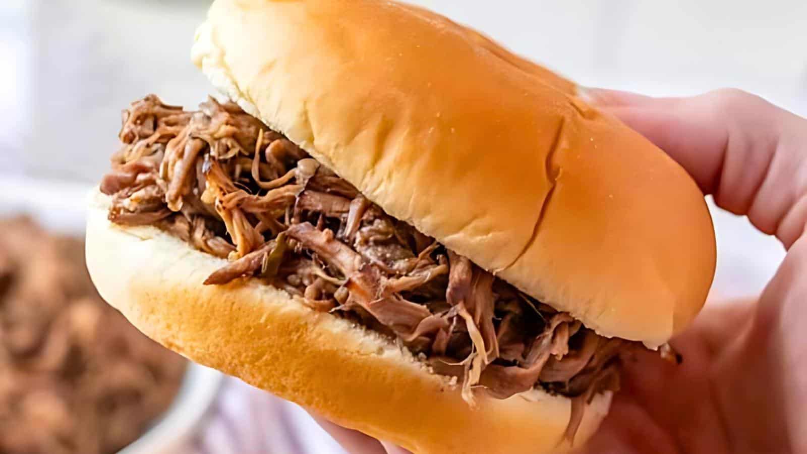 A hand holding a pulled pork sandwich in a soft, golden bun. The shredded meat is generously piled between the halves of the bun, with a slightly blurred bowl of pulled pork in the background.