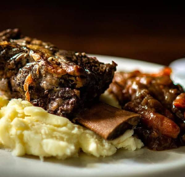 A serving of braised short ribs on mashed potatoes with stewed carrots and beans on a white plate.