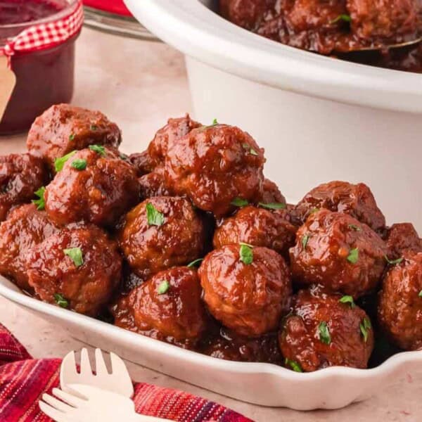 A white dish with glazed meatballs garnished with herbs, near fresh cranberries, parsley, a red cloth, and a slow cooker.