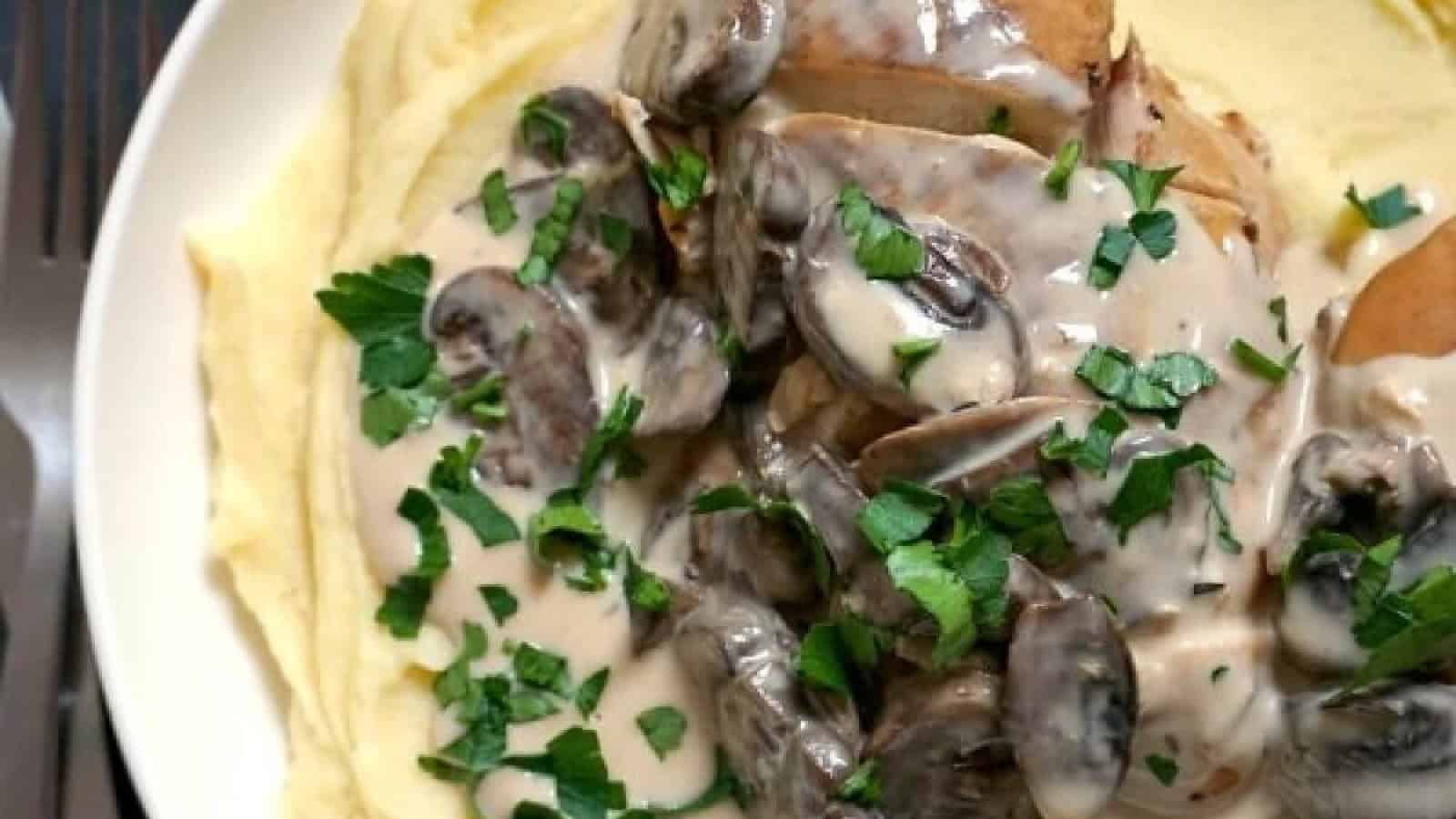 A plate of creamy mashed potatoes topped with a serving of chicken and mushroom sauce, garnished with chopped fresh parsley. The mushrooms are sliced and mixed in a rich, creamy sauce, adding to the hearty appearance of the dish. A fork is placed to the side.