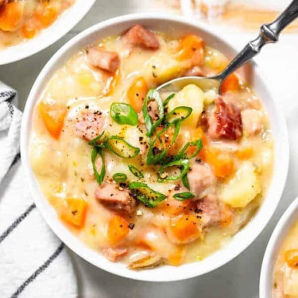 A bowl of creamy soup containing diced ham, carrots, potatoes, and green onions with a spoon on the side.