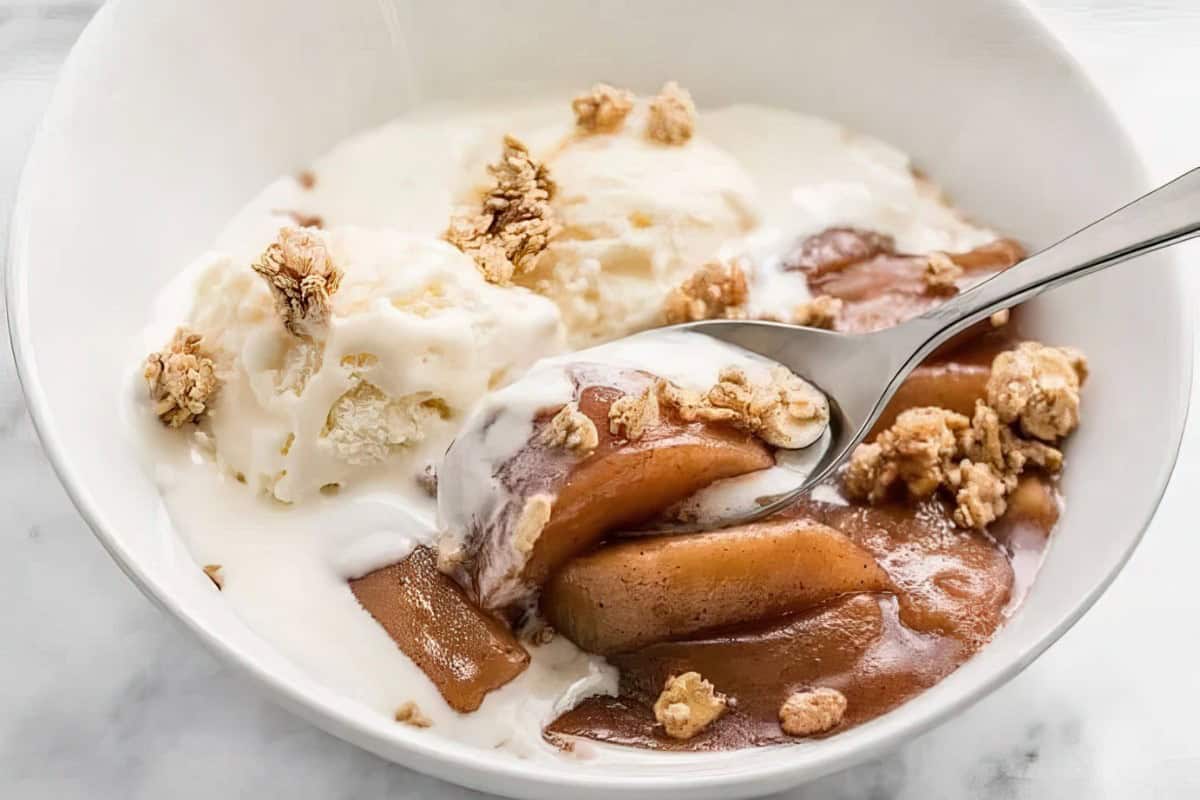 A white bowl contains a dessert with creamy vanilla ice cream, tender baked apple slices coated in a cinnamon sauce, and a sprinkle of crunchy granola on top. A spoon is scooping up a portion of the dessert, capturing a mix of all the elements.