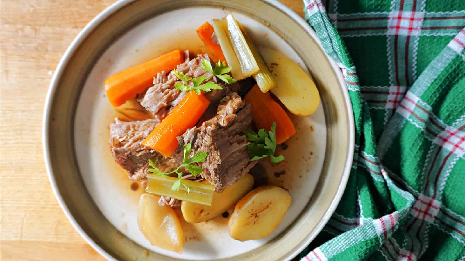A plate of pot roast with tender beef chunks, carrots, celery, and potatoes, garnished with fresh herbs, sits on a wooden surface next to a green and red plaid cloth. The dish appears hearty and flavorful, served with a bit of broth.