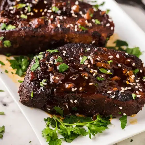 Two grilled, glazed steaks with sesame seeds and chopped herbs on a white plate with fresh parsley garnish.