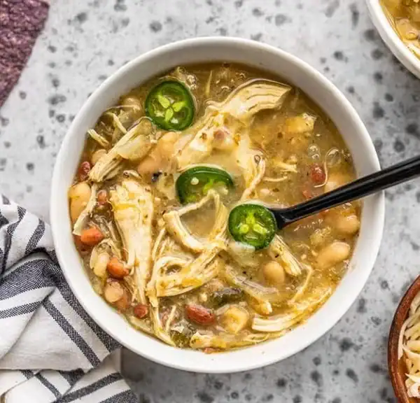 A bowl of white chicken chili with shredded chicken, white beans, sliced jalapeños, and a black spoon on a gray surface next to a striped napkin and a small bowl of shredded cheese.