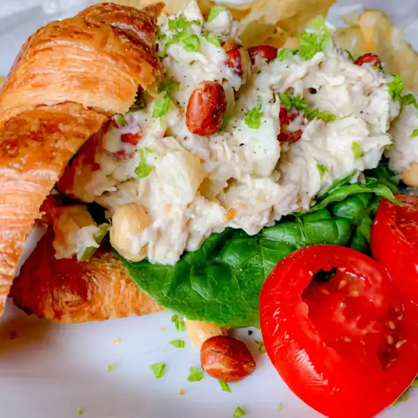 A croissant sandwich with chicken salad, lettuce, and sliced tomatoes on a white plate. Chips are on the side.