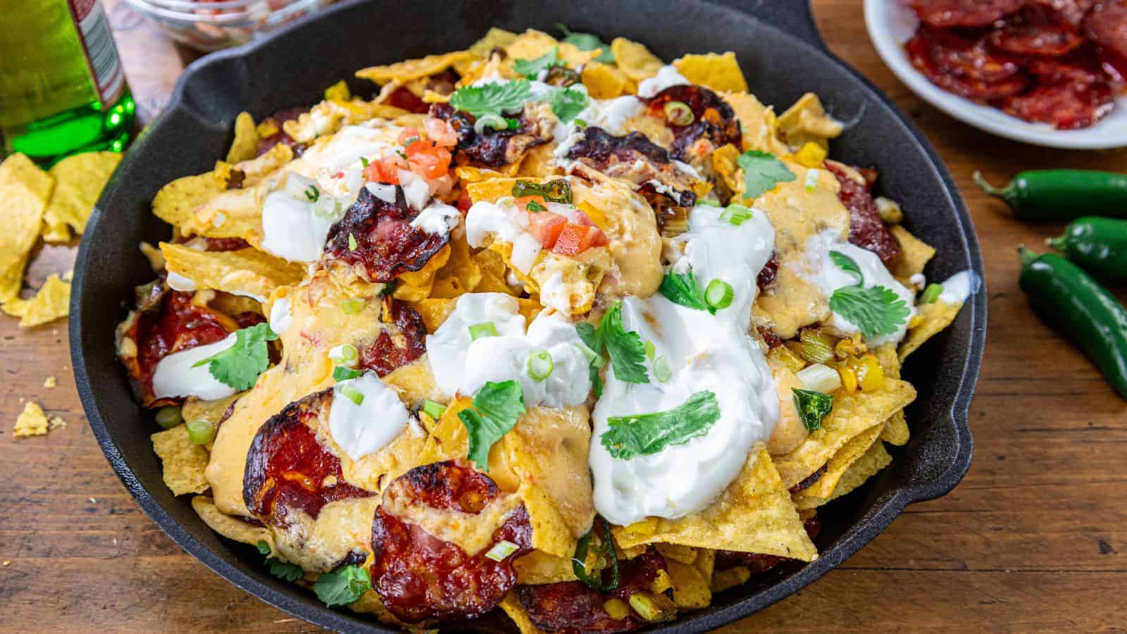A skillet of nachos topped with melted cheese, sour cream, sliced green onions, tomatoes, cilantro, and pepperoni slices.