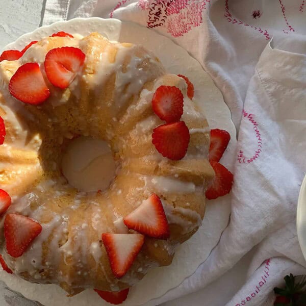 A Bundt cake glazed and topped with sliced strawberries, surrounded by bowls of whole strawberries on a white surface with a patterned cloth.