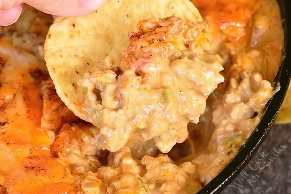 Close-up of a hand holding a tortilla chip, dipping it into a creamy, cheesy chicken and vegetable mixture. The dip is vibrant and gooey, with visible chunks of chicken and melted cheese, giving it a rich and appetizing appearance.