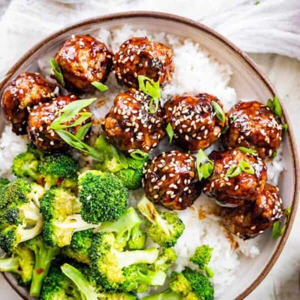 A bowl containing white rice, broccoli florets, and glazed meatballs garnished with sesame seeds and chopped green onions.