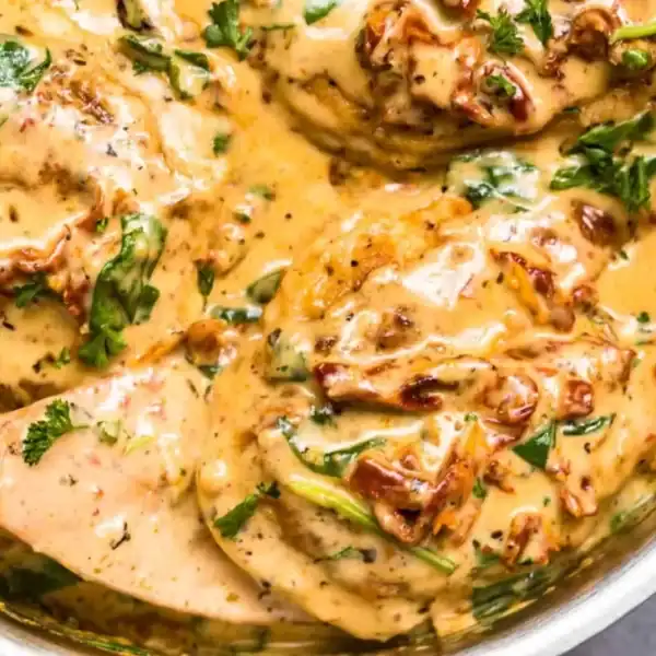 Chicken breasts in creamy sauce, sun-dried tomatoes, and fresh herbs in a pan with a wooden spoon.