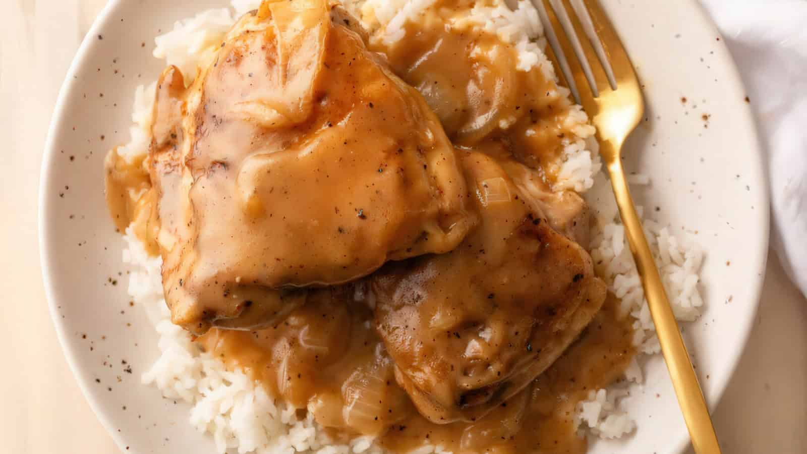 A plate of rice topped with two pieces of cooked chicken covered in a thick, brown gravy. A gold fork is placed on the right side of the plate.