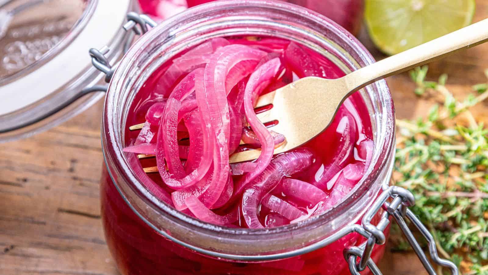 A glass jar filled with pickled red onions, with a gold fork lifting some onions out.