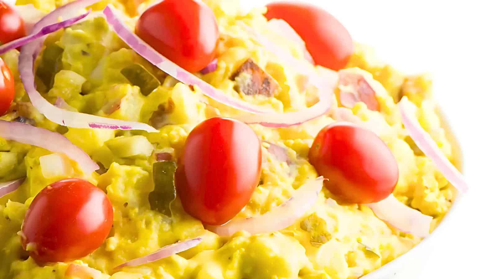 A close-up of a vibrant dish of potato salad topped with halved cherry tomatoes and thin slices of red onion. The potato salad appears creamy and is flecked with green herbs and pickles. The image has a bright and fresh appearance.