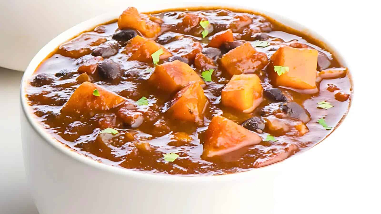 A white bowl filled with a hearty, chunky stew featuring pieces of sweet potato, black beans, tomatoes, and other vegetables. The stew has a thick, reddish broth and is garnished with small green herbs.