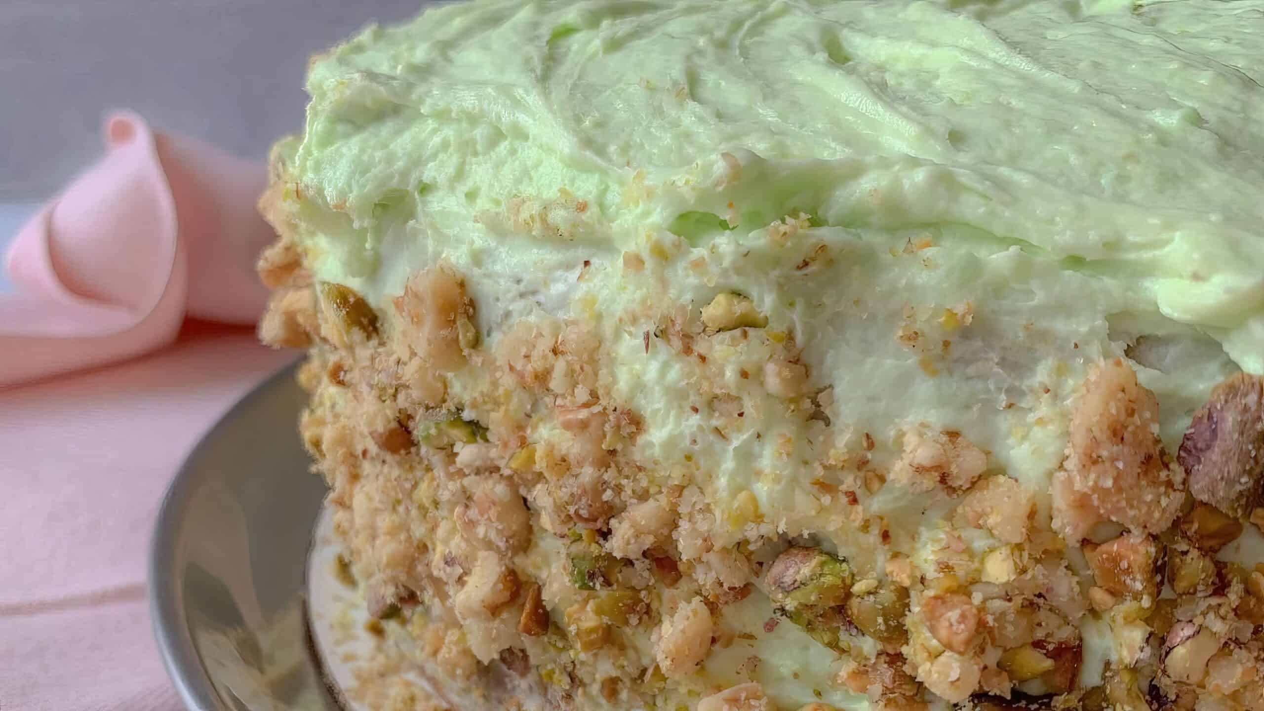 Close-up of a cake with light green frosting and a garnish of crushed nuts on the sides. The cake is placed on a silver plate, and a folded pink napkin is visible in the background.