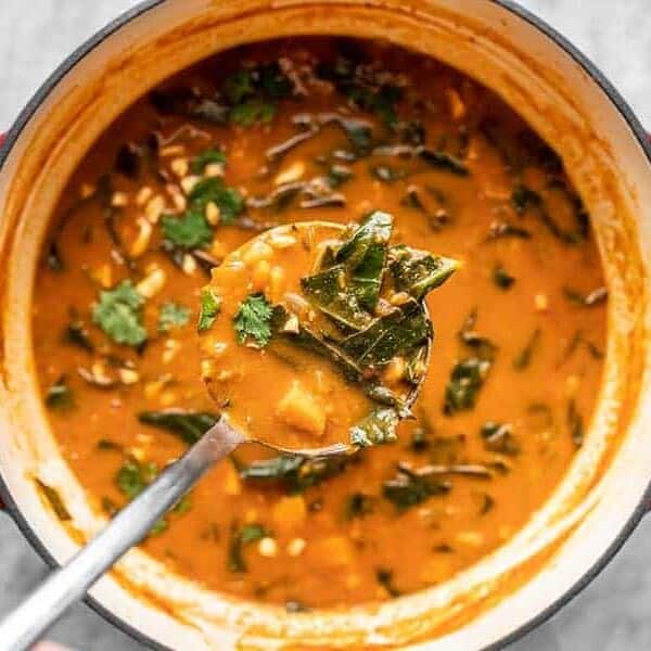 A pot of vibrant, reddish-orange soup containing pieces of greens, vegetables, and herbs. A metal ladle is lifting a spoonful of the soup, which shows chunks of vegetables and leafy greens, over the pot with large red handles, placed on a light grey surface.