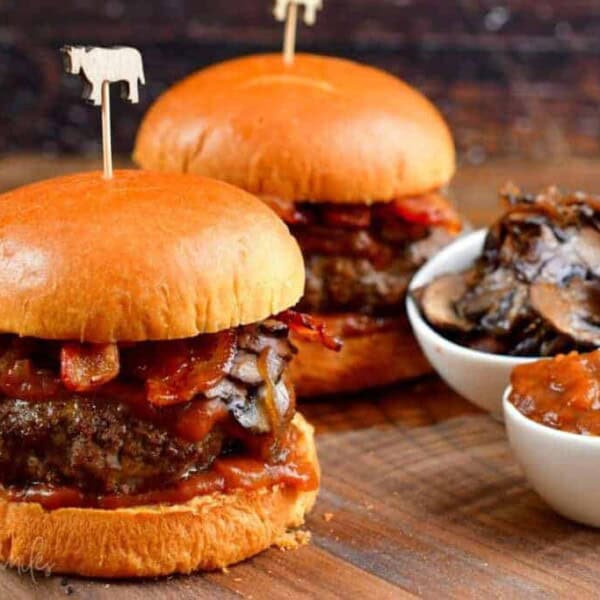 Two juicy burgers with melted cheese, crispy bacon, and sautéed mushrooms are served on toasted buns. Each burger is held together with a skewer topped with a cow-shaped decoration. There are small dishes of sautéed mushrooms and smoky barbecue sauce on the side.