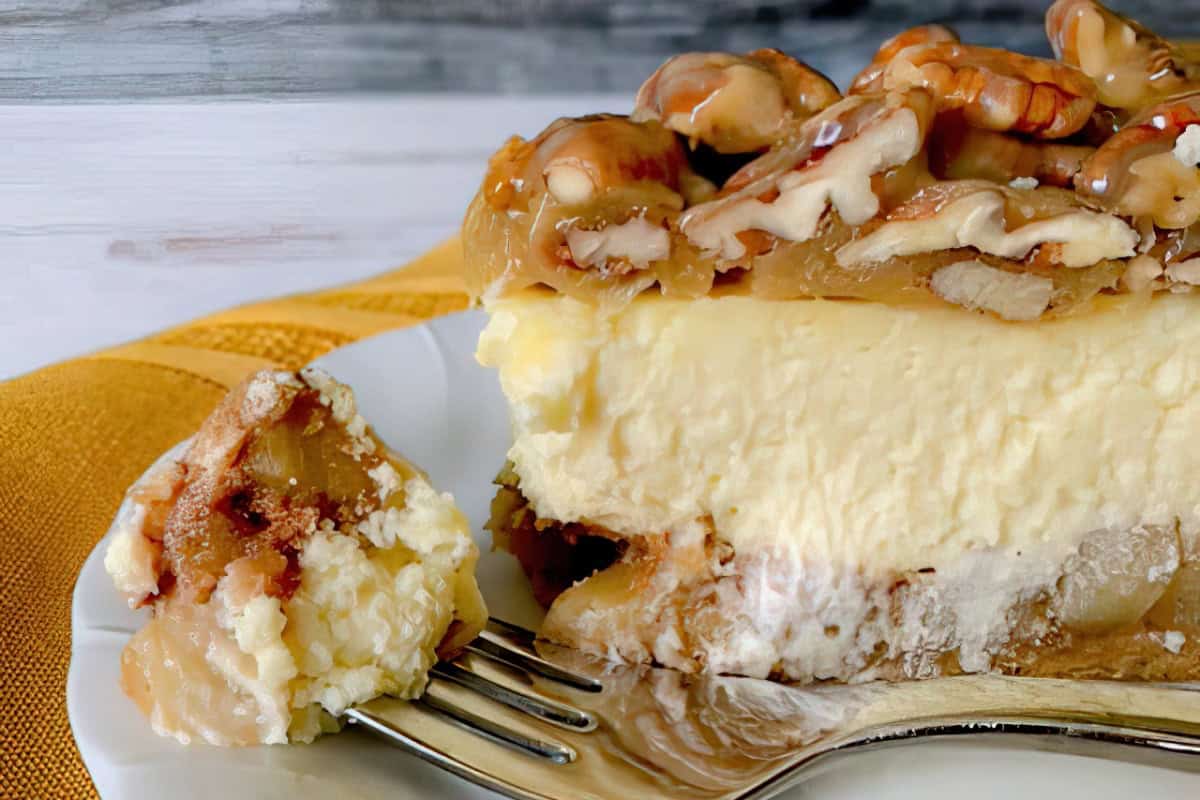 Close-up of a slice of creamy cheesecake topped with glazed pecans on a white plate. A fork with a small piece of cake is placed in front, resting on a yellow cloth. The background features a light, textured surface.