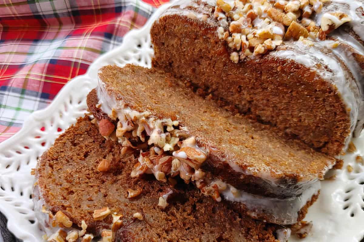 Close-up image of sliced banana bread topped with chopped nuts and a glaze, placed on a white, intricately designed plate. The background features a red and green plaid cloth. The focus is on the mouth-watering texture and toppings of the bread.