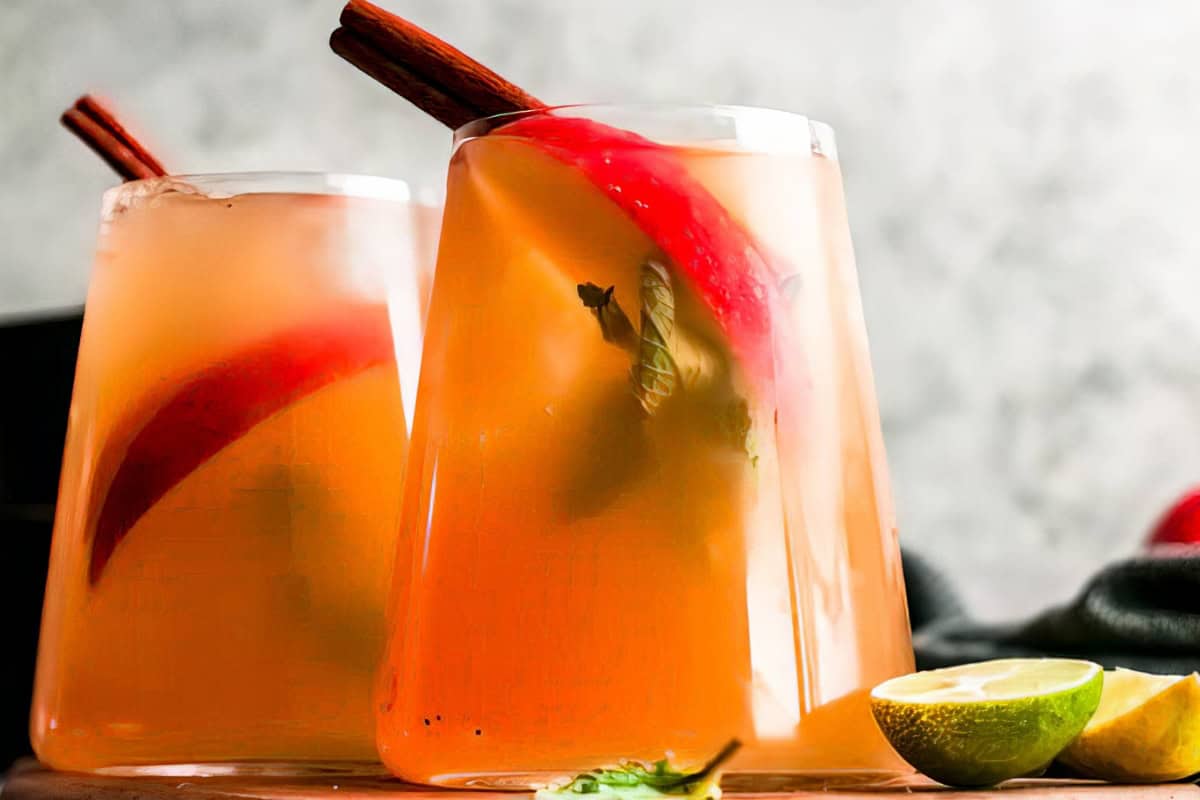 Two glasses of cold, orange-hued punch with red apple slices and cinnamon sticks garnish the drinks. Fresh lime and lemon wedges are scattered at the base of the glasses. The background is out of focus, creating a warm and inviting scene.