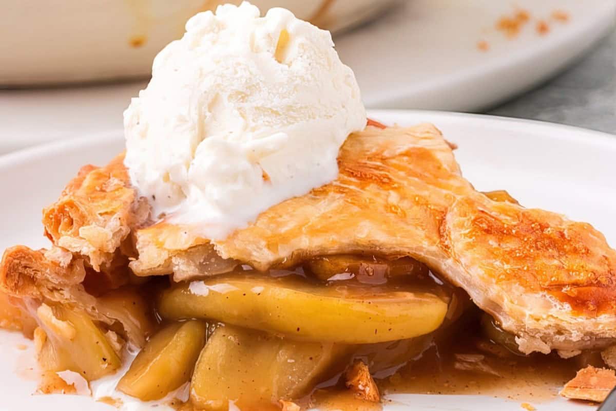 A slice of apple pie on a white plate, topped with a scoop of vanilla ice cream. The golden-brown crust is flaky, and the filling includes tender, caramelized apple slices. The background shows a glimpse of a pie dish.