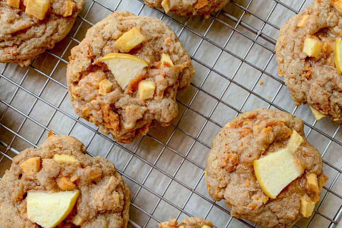 Close-up of freshly baked apple-cinnamon cookies on a cooling rack, featuring visible apple chunks and a golden-brown, slightly crumbly texture.