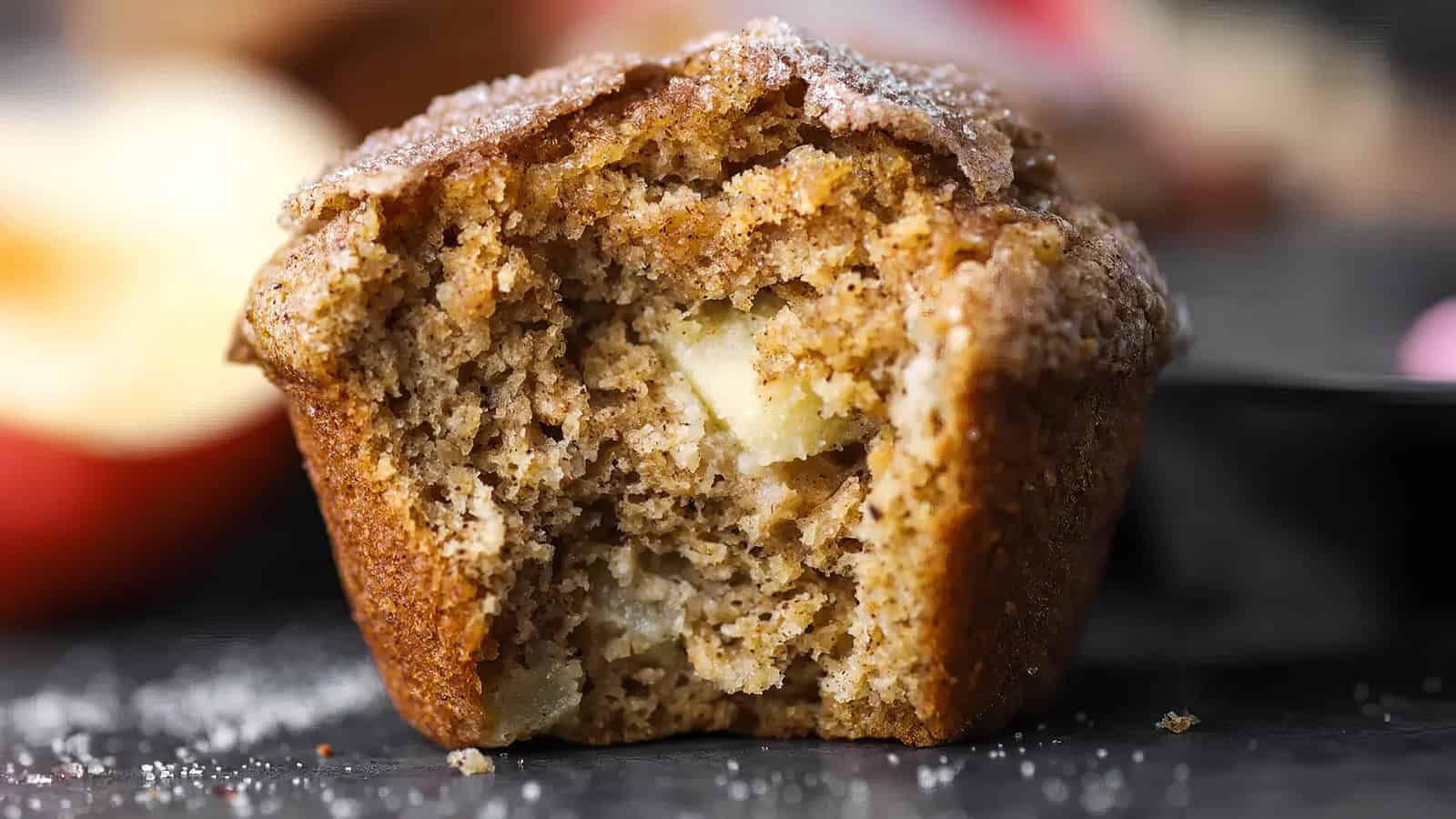 Close-up of a partially eaten muffin with visible apple pieces, showcasing a soft, moist interior. The golden-brown muffin is topped with a dusting of powdered sugar, adding to the appetizing appearance. The background is a blurred dark surface with hints of an apple.