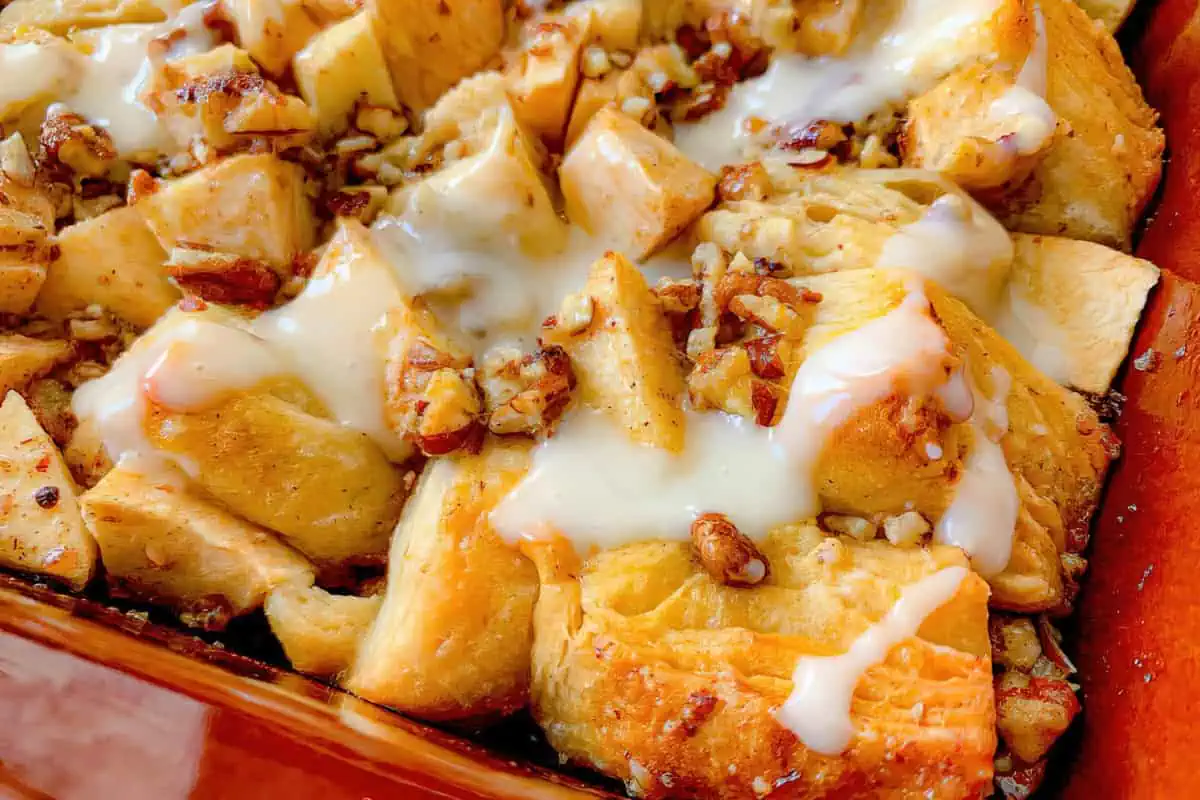 A close-up of a baked dish featuring golden-brown dough pieces topped with chopped nuts, small chunks of apple, and drizzled with a creamy white glaze. The texture appears soft and slightly crisp, and the dish is presented in a reddish-brown baking tray.